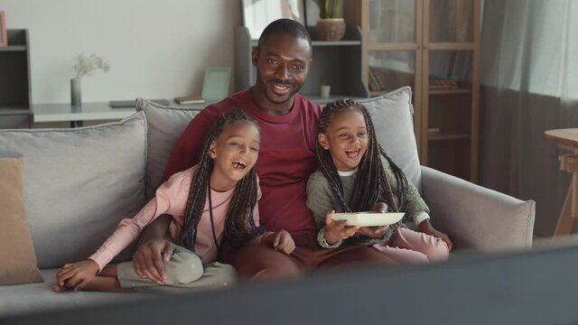 High angle of young African man and two adorable female kids wearing braids sitting on couch at home, watching TV, laughing, having snacks