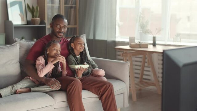 Steadicam of happy African man and two adorable female kids wearing braids sitting on couch at home, watching comedy on TV, having snacks in evening