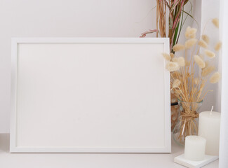 Mock up poster frame  dried Lagurus ovatus flowers composition in modern glass vase and candles on  white table and cement wall background