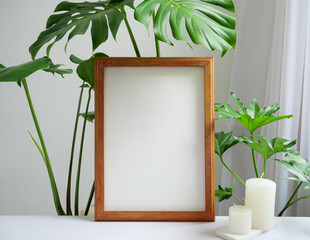 Mock up brown poster frame, gouse plant Philodendron green leaf and aroma white candles on beige...
