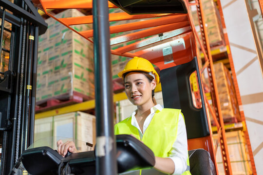 Forklift driver asian young woman in safety jumpsuit uniform with yellow hardhat at warehouse. Worker female  working and smiling in forklift loader work