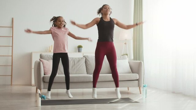 Intensive cardio training. Energetical african american mother and daughter practicing Jumping Jack exercise, at home