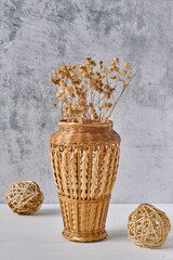 Brown knitted vase made of twigs for weaving with dried flower and rattan balls on white wooden table against gray cement background. Concept of handmade, comfort and eco decor. Home interior.