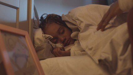 Low-angle of adorable little African girl sleeping in bed, hand of unrecognizable parent covering her with blanket and patting on head