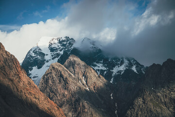 Obraz premium Scenic landscape with great rocks and snowy mountains in sunlight in low clouds. Wonderful view to mountain top with snow in sunshine in cloudy sky. Awesome scenery with sunlit snow-white pinnacle.