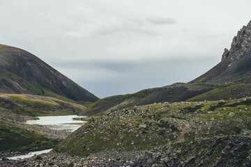 Atmospheric mountain landscape with mountain lake near sharp rocks in narrow valley in rainy weather. Bleak overcast scenery with mountain lake among rocky hills in highland glen under gray cloudy sky