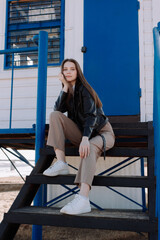 Smiling young woman in black leather jacket sitting on stairs of watchtower in front of blue door. Having fun outside. Urban style and street fashion. Girl in casual outfit looking at camera