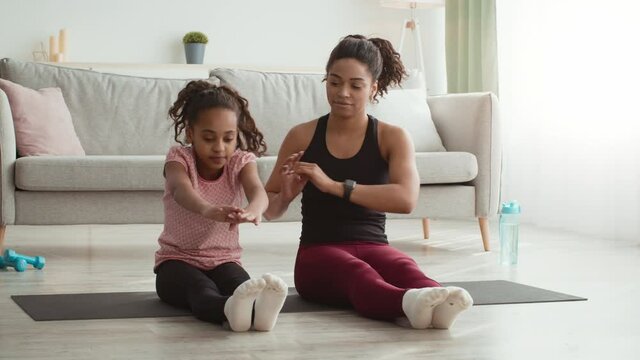 Pilates training. Young flexible black mother helping her daughter to stretch, practicing body stretching exercise