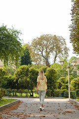 Fototapeta na wymiar A beautiful woman with blond hair walks through the cozy green park of the city. Summer mood. Walk around a European city on a weekend. Beautiful historic architecture. Travel to Europe, tourism.