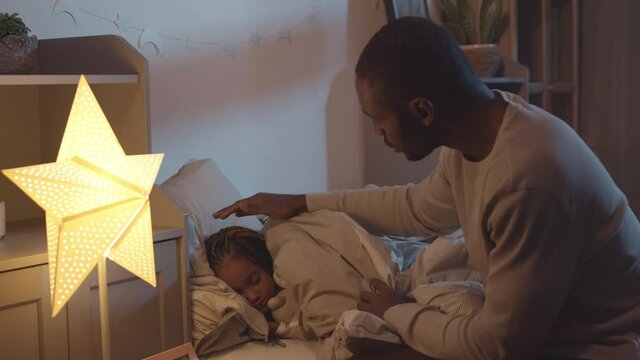 Side view medium long of young African father sitting on edge of bed in kids room, tacking in blanket for his little daughters, patting one on head, then leaving frame