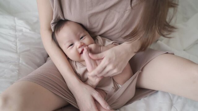 asian mother parent playing with baby infant enjoy sucking hand fingers and foot. kid laughing when play with mom on bed in bedroom. life at home. happy newborn baby learning development skill lesson.