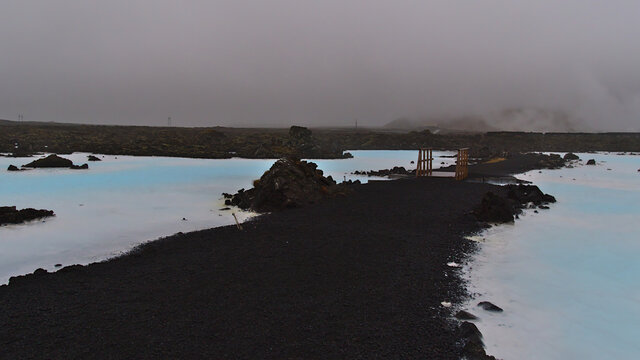 View of wooden footbridge crossing pools of blue shimmering water in a lava field near geothermal spa resort Blue Lagoon, Grindavik, Reyjanes peninsula, Iceland on cloudy day in winter season. - Powered by Adobe