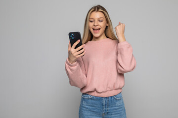 Portrait of a young woman holding mobile phone and celebrating over gray background