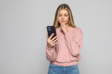 Naklejka premium Portrait of pretty woman in shirt, denim outfit holding hand in pocket of jeans, having smart phone, checking email, searching, using 3G internet, standing on grey background