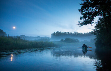 Hikers rowing an canoe in the summer night river, surrounded by fog clad floodplain and under full moon in the sky on the Halliste river in Soomaa NP
