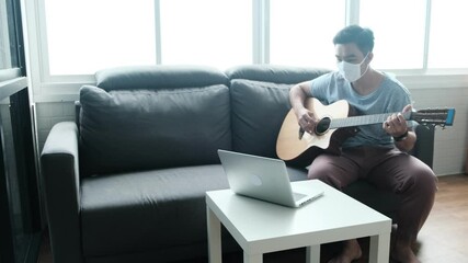 Musician Asian male playing acoustic guitar instrument singing song learning practicing carefree time spend activity occupation sitting on sofa in living room quarantine lockdown staying safe at home