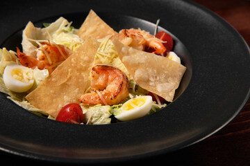 Appetizing Caesar salad with fried shrimps in a black plate on a dark wooden background. Close-up of a dish. Healthy and dietary food.
