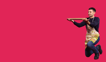 Brunette man, caucasian wearing chef apron kneeling holding rolling pin, looking at empty space for your message info ad, full length on red background with copy space. Fun in the kitchen