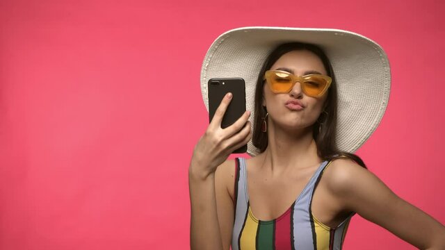 Woman in sun hat and sunglasses taking selfie isolated on pink