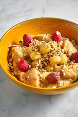 Tasty and healthy oatmeal with honey, apple, raspberry and sunflower seeds in a bright yellow bowl on a white marble background.