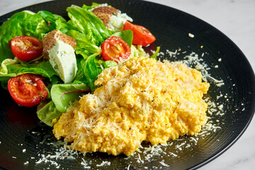Scrambled omelet with spinach salad, ricotta cheese and cherry tomatoes, served in a black plate on a marble background. Delicious breakfast
