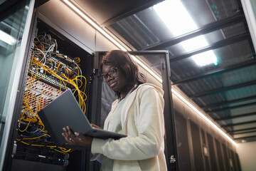 Low angle portrait of African-American female network engineer standing by server cabinet and...