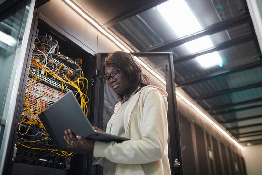 Low angle portrait of African-American female network engineer standing by server cabinet and holding laptop while working with supercomputer in data center, copy space