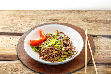Soba with mushrooms and sesame seeds in a plate on a stand on a wooden table next to chopsticks.