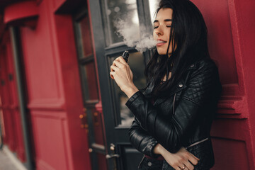 Obraz na płótnie Canvas Beautiful stylish brunette smoking an e-cigarette as she is walkind through the city. Vaping concept. Trendy new vaping device, smoke e-liquid instead of nicotine cigarettes.