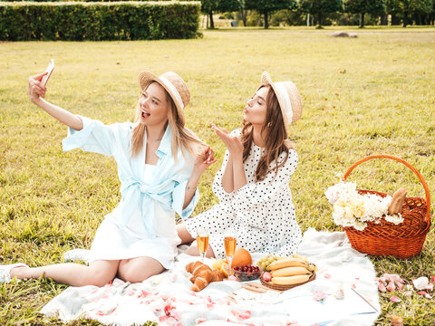 Two young beautiful hipster woman in trendy summer sundress and hats. Carefree women making picnic outside. Positive models sitting on plaid on grass, eating fruits and cheese.Taking selfie photos