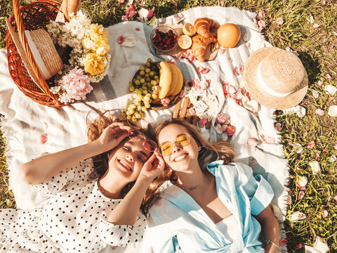 Two young beautiful smiling female in trendy summer sundress and hats.Carefree women making picnic outside.Positive models sitting on plaid on grass, eating fruits and cheese. Top view