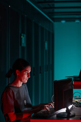 Vertical side view portrait of female IT engineer using computer while working in server room lit by blue light, copy space