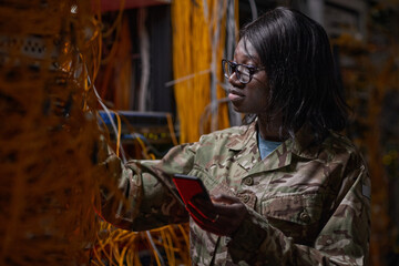 Side view portrait of young African-American woman wearing military uniform inspecting server with cables and wires