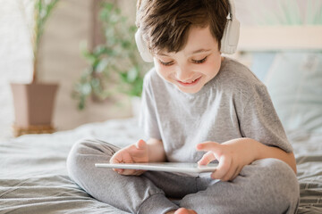 Happy kid lying on the bed at home and playing with pc tablet or reading online and listening to...