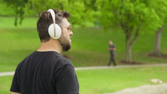 Back view of handsome young man listening music in modern wireless white headphones. Smiling cheerful man shaking head to the beat in green urban park at summer sunny day. RED Weapon Helium 8K footage