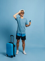 Happy caucasian male tourist, in a hat and sunglasses, looks into the distance to meet adventures, next to him a blue suitcase, rejoices a long-awaited vacation, stands on an isolated blue background