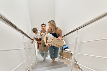 Happy family couple new home owners real estate buyers or renters with toddler kid child son carrying boxes going up stairs on moving day arriving into new apartment. Relocation, mortgage, removals.