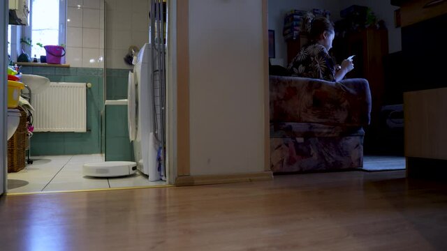 Robotic vacuum and mop cleaner cleans around furniture indoors and changes direction of moving automatically using it's sensor while a female is sitting on a couch with cellphone.