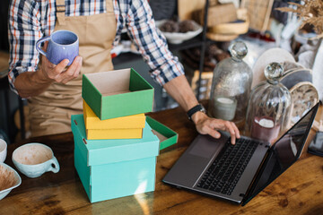 Close up of store worker rechecking available assortment and using modern laptop. Counter with various beautiful decor. Competent seller at workplace.