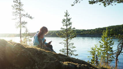 Happy curly hair mother with little daughter sitting by lake during sunset on spring camping vacation and browsing internet on mobile phone device watching video. Concept of nature quality family time