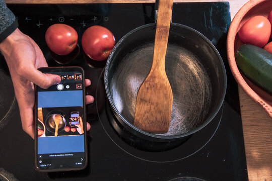 hand holding mobile with the same picture two pieces of red tomato next to ceramic hob with a black pot and wooden spoon