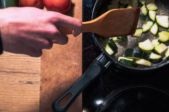 a hand cooking and two pieces of red tomato next to ceramic hob with a vegetable on a black pot on wooden background