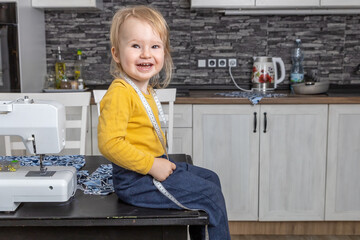 Cute laughing little boy is sitting on the table with tailor’s tape measure  smiling at the camera.