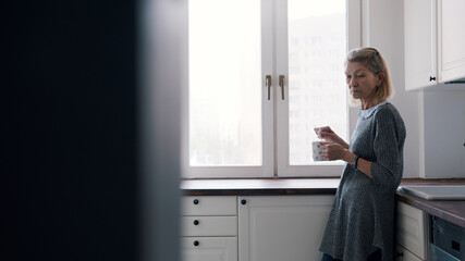 Depressed lonely elderly woman mixing sugar in the hot beverage while standing in her kitchen. High quality photo