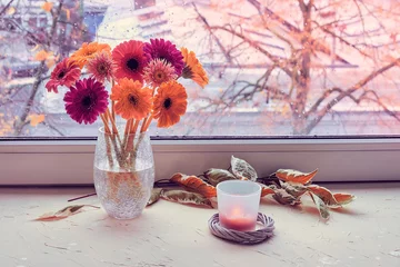 Fotobehang Autumn window, gerbera flowers, candle. Pink, magenta, coral and yellow Gerbera flowers on a window board. Green white leaves. Grey cold weather outside, orange sunset. Stay warm and cozy indoors. © tilialucida