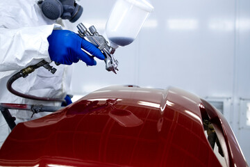 Car painter in protective clothes and mask painting automobile bumper with metallic paint and...