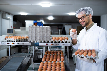 Portrait of food factory worker with hairnet and hygienic gloves checking quality of eggs by...