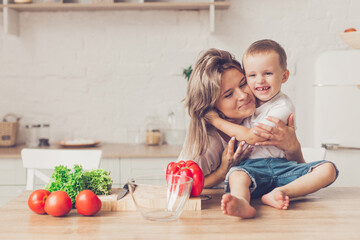 Son with mom are cutting vegetables on salad in kitchen and hugging. Happy family.