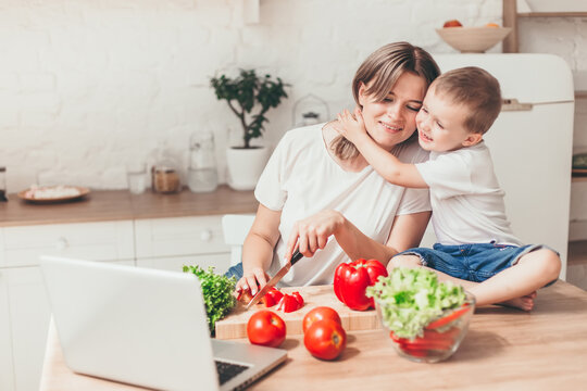 Son With Mom Are Cutting Vegetables On Salad In Kitchen And Hugging. Happy Family.