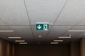 green emergency exit sign on a white ceiling with neon light on, with fire alarm in blurred background, by day, without people